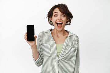 Portrait of happy and surprised cute girl showing her phone screen and looking at camera in awe, recommending download an app, standing over white background