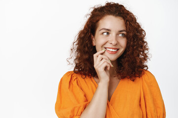 Fototapeta na wymiar Women wellbeing. Attractive redhead woman with curly hairstyle smiling and looking thoughtful aside, thinking with pleased smile, standing in orange dress over white background