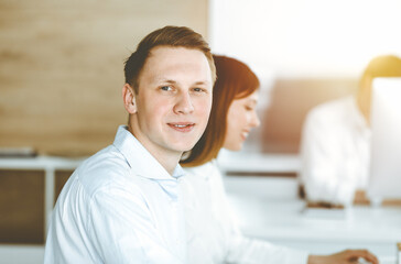 Obraz premium Cheerful smiling businessman at work in sunny office. Casual dressed entrepreneur using pc computer while sitting with diverse colleagues at the background