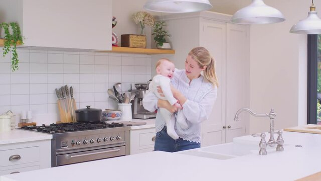 Loving mother carrying laughing baby son around kitchen at home playing game together