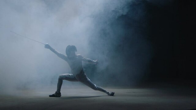 Professional fencer in protective uniform practicing maneuvers with sword alone in dark studio . Fencing demonstration on black smoked background . Shot on Arri Alexa cinema camera in slow motion .	