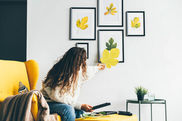Young woman with long loose curly hair puts dried leaves applique on frame glass and covers with sheet of paper against decorated wall at home low angle shot - Powered by Adobe