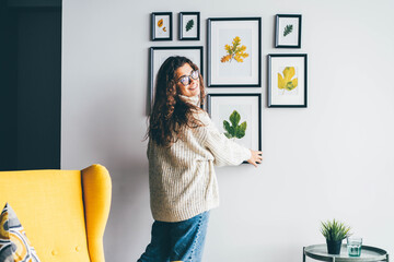 Skilled woman designer with long loose curly hair holds wooden frame with dried leaf applique and hangs on decorated wall in new apartment backside view. - obrazy, fototapety, plakaty