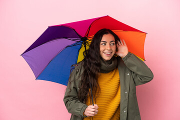 Young woman holding an umbrella isolated on pink background listening to something by putting hand on the ear