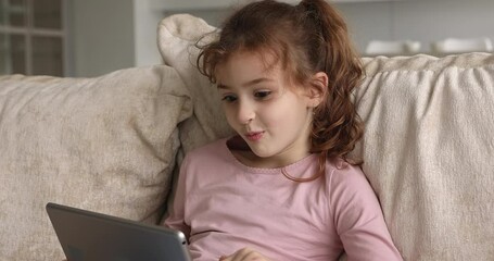 Happy adorable small child girl holding digital computer tablet in hands, playing online games, having fun communicating distantly in social network or web surfing, modern technology addiction concept
