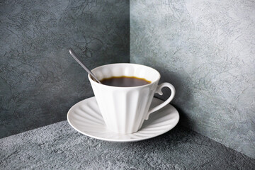 White porcelain hot coffee cup with spoon and saucer on gray surface in the corner.