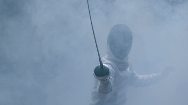 Professional fencer in protective uniform practicing with sword in dark studio . Fencing demonstration on black smoky background . Shot on Arri Alexa movie camera in slow motion