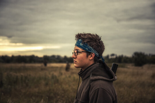 Hunter teenager boy portrait standing alone in rural field at dramatic sunrise during hunting and looking into the distance  