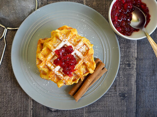 gluten-free pumpkin Belgian waffles sprinkled with powdered sugar and currant jam lie with cinnamon sticks on a gray plate on a wooden table top view