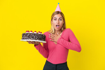 Blonde Uruguayan girl holding birthday cake isolated on yellow background surprised and shocked while looking right