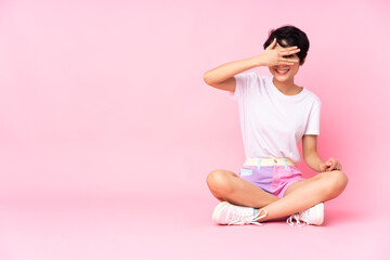 Young Vietnamese woman with short hair sitting on the floor over isolated pink background covering eyes by hands and smiling