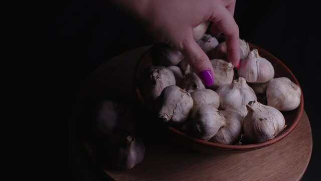 Pile of whole bulbs of garlic in ceramic bowl on table. Woman's hand takes garlic from bowl