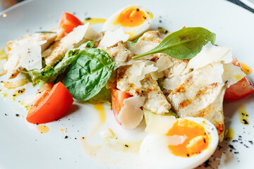 Classical Caesar salad with roasted chiken, eggs, lettuce leaves, parmesan and cherry tomatoes on the white plate close up