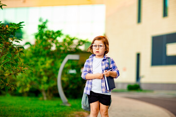 a little girl, a schoolgirl, wearing glasses, is standing near the school, with a backpack, holding...