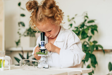 Little girl in white uniform and protective glasses looks into the microscope in a home...