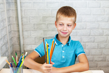 A smiling child is sitting at a desk and holding a bouquet of pencils in his hands. Office supplies are laid out on the table. Back to school.