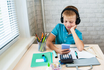 Smiling boy is sitting at desk with headphones on and holding mobile phone in his hands. Office supplies are laid out on table. Back to school. Holidays.
