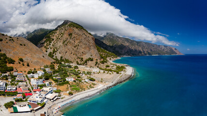 Panoramic aerial view of a small coastal village with towering, cloud covered mountains (Agia...
