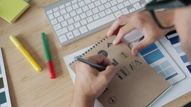 Close-up of designer's hand drawing logos in notepad working at desk in office while creative specialist is concentrated on designing logotypes