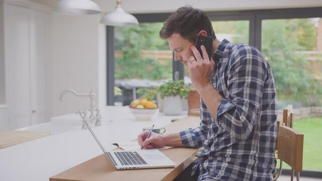 Man working from home using laptop on kitchen counter talking on mobile phone- shot in slow motion