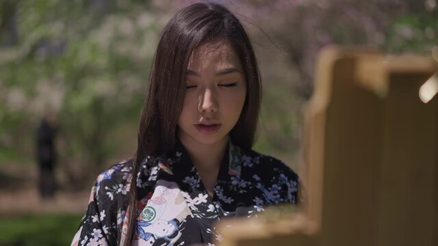 Portrait of confident concentrated Asian woman looking at paint brush and drawing on easel in slow motion standing in sunny spring park. Artist painting nature outdoors on sunny day