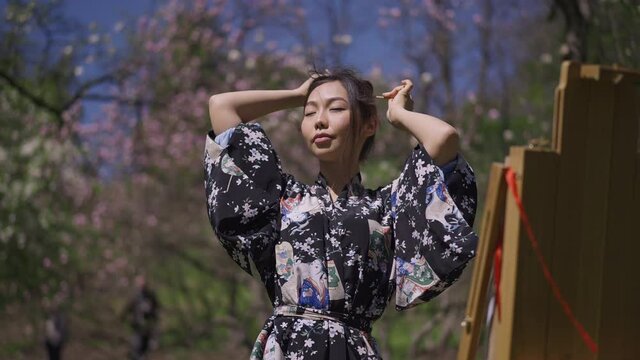 Medium shot portrait of young creative Japanese woman in kimono making ponytail looking at easel in blooming spring park. Confident smiling beautiful slim artist painting outdoors in sunlight