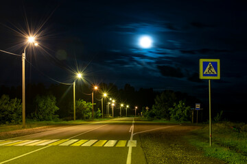 a mysterious, gloomy view of an asphalt road with a pedestrian crossing, road signs, bright street lights with a bus stop against the background of the night sky in a full moon