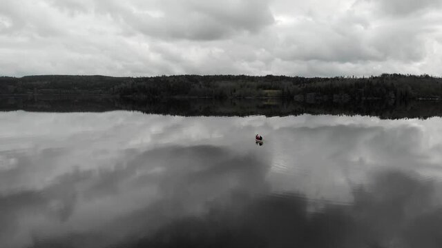 Dolly in descending aerial shot of two people paddling gently on a white canoe while creating small ripples all over a calm reflective lake towards land on a grey cloudy day.