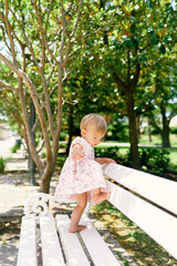 Little barefoot girl in a dress climbs on a park bench