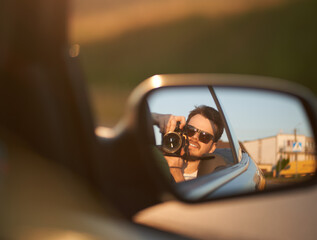 Fototapeta na wymiar Selfie of happy man with camera in convertible's car side view mirror. Concept of freedom and vacation adventures