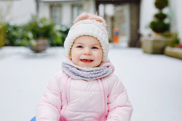 Cute little toddler girl enjoying a sleigh ride on snow. Child sledding. Baby kid riding a sledge in colorful fashion clothes. Outdoor active fun for family winter vacation on day with snowfall