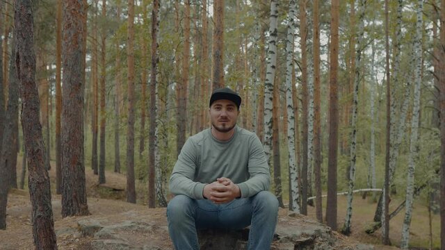 A Man in a Black Cap Sits in the Forest Among the Trees and Looks to the Parties. Lost Man in the Forest. Tourist Enjoys the Beauty of the Forest. Hicker in the Woods at Rest