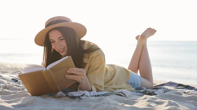 Excited traveler tourist young brunette woman 20s wear summer casual clothes straw hat read book lying on plaid have picnic isolated on sea beach background outdoors. People vacation lifestyle concept