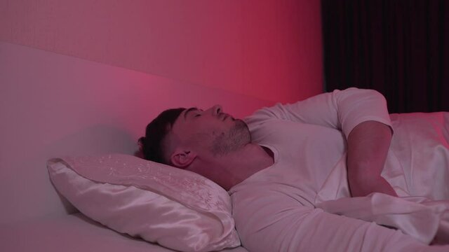 Young Tired Man Sleeping in Bed in His Bedroom. He Smiles in Sleep and Continues to Rest. The Room Is Lit Red From A Night Lamp. Cozy Sleep at Your Home. Sleep and Rest concept