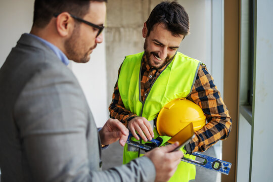 Smiling construction worker standing with supervisor and looking blueprints on tablet while standing in building in construction process.