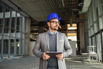 Supervisor standing in building in construction process, holding tablet and checking on works.