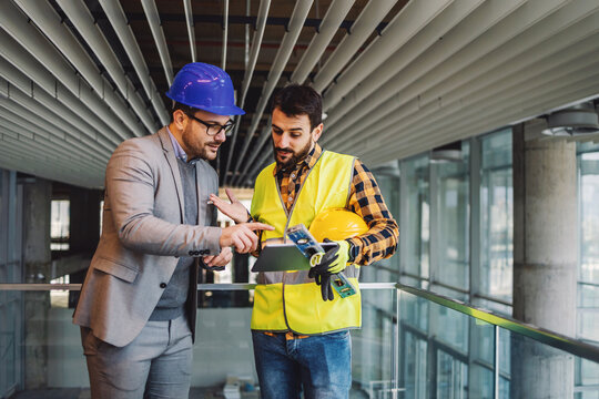 Architect And Construction Worker Standing In Building In Construction Process And Looking At Blueprints On Tablet.