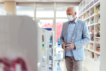 Naklejka na ściany i meble Shopping of drugs in pharmacies and drugstores and corona virus. A mature man wearing a suit and a protective face mask standing in the middle of a shelf with medicines and looking at the drugs shelf