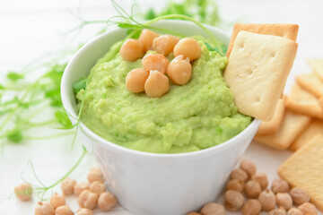 Bowl with tasty green pea hummus, chickpeas and crackers on light background, closeup