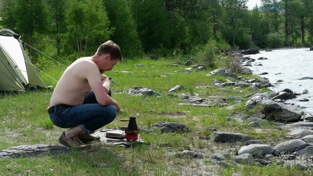 Fifty-year-old blond man brews coffee on rocky bank of amountain river. Slow motion