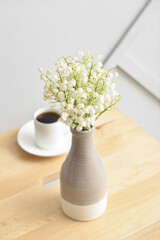 Vase with lily-of-the-valley flowers and cup of coffee on table near light wall
