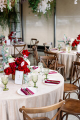 Decoration of the wedding table with red and pink flowers on a white tablecloth. Red glass goblets and burgundy napkins on the plates.