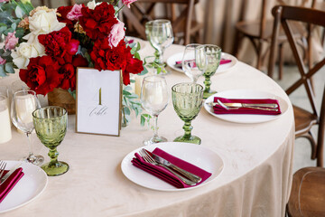 Decoration of the wedding table with red and pink flowers on a white tablecloth. Green glass goblets and framed table number.