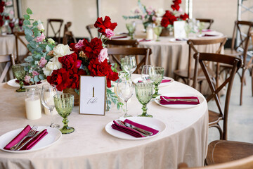 Decoration of the wedding table with red and pink flowers on a white tablecloth. Green glass goblets and framed table number.