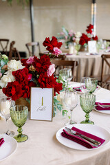 Decoration of the wedding table with red and pink flowers on a white tablecloth. Green glass goblets and framed table number.