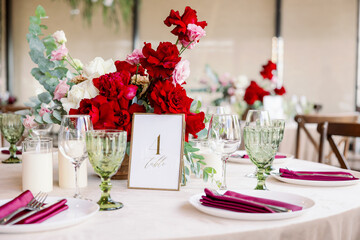 Decoration of the wedding table with red and pink flowers on a white tablecloth. Green glass goblets and framed table number.
