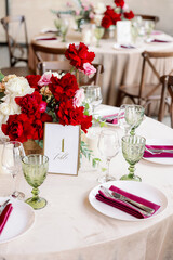 Decoration of the wedding table with red and pink flowers on a white tablecloth. Green glass goblets and framed table number.