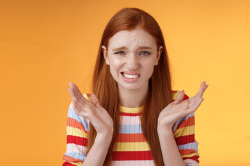 Awkward unhappy worried young redhead girl cringe feel sorry apologizing smirking smiling nervously frowning squinting spread hands sideways shrugging confused, standing orange background - obrazy, fototapety, plakaty