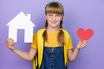 Portrait of beautiful cute smiling schoolgirl kid holding small red heart and paper white house model, happy looking at camera, wears yellow backpack, isolated over pastel purple background in studio