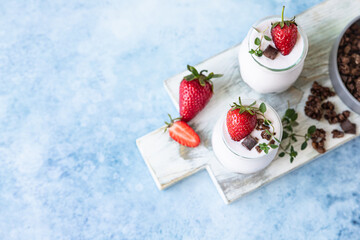 Two jars with yoghurt, chocolate granola and fresh strawberries on ceramic plate, blue concrete background. Top view.